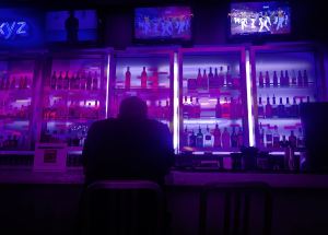 image of a man sitting at a bar, looking at TV above bar with neon lit bottles behind bar.